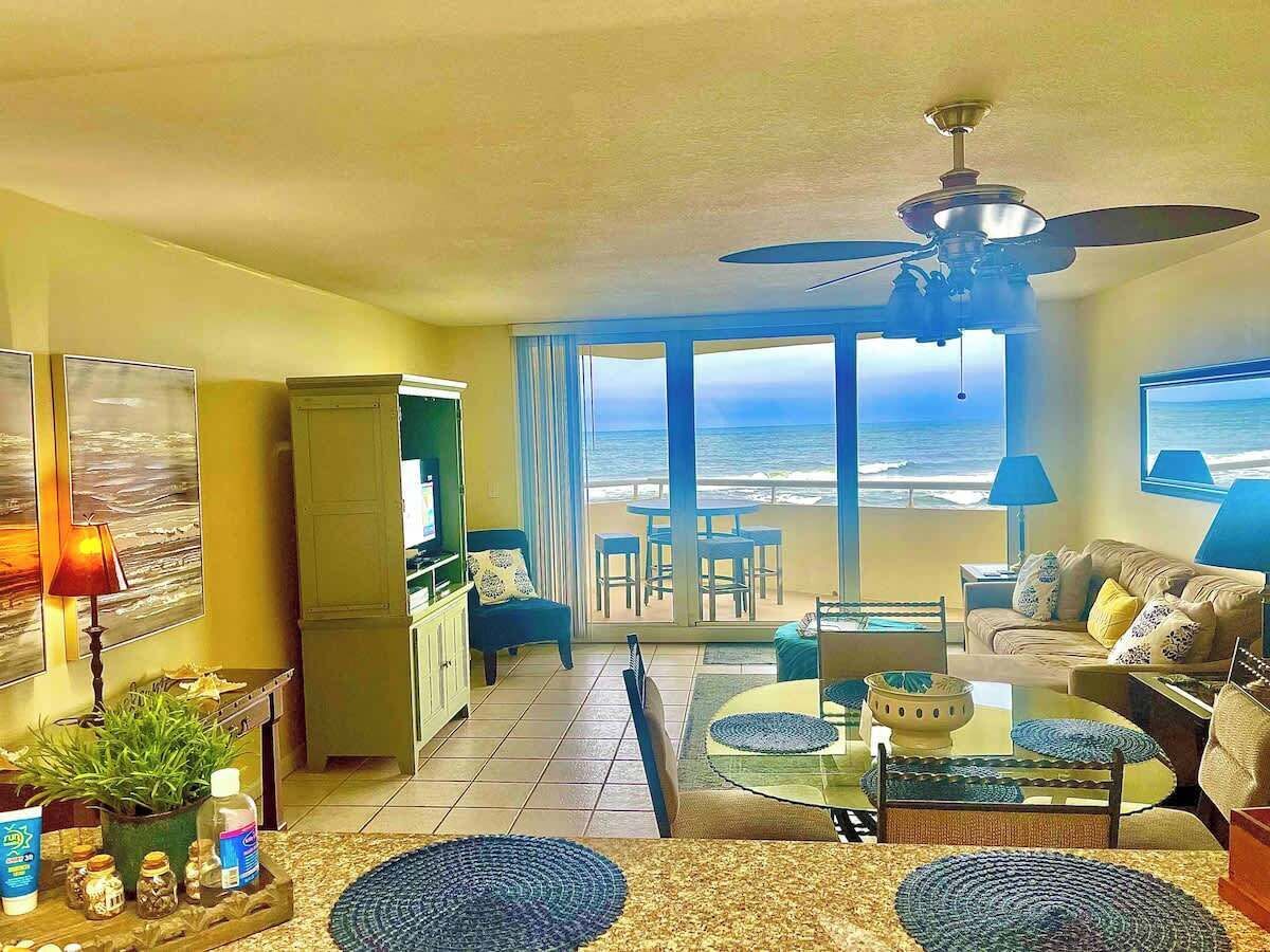 A living room with a view of the ocean, a ceiling fan, and a dining area with a table and chairs.