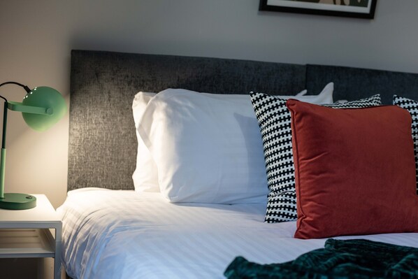 A neatly made bed with white and patterned pillows, a green floor lamp, and a grey headboard.