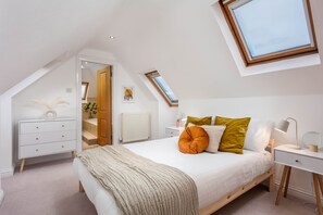 A bedroom with a sloped ceiling, a skylight, a bed with pillows, a bedside table with a lamp, and a dresser.