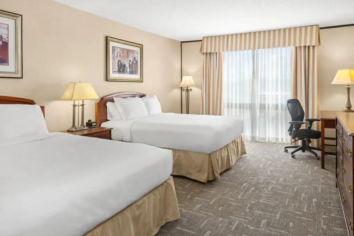 Habitación de hotel con dos camas, un escritorio, una silla y un ventanal con cortinas.