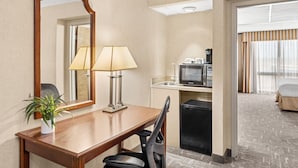 A hotel room with a wooden desk, a lamp, a mirror, a small kitchenette, a television, a bed, and a potted plant.