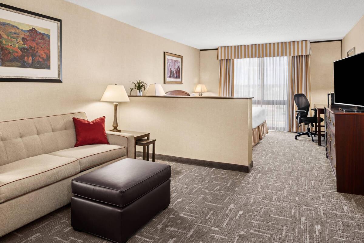 Habitación de hotel con sofá, reposapiés, escritorio, lámpara, silla, televisor y cama con cortinas.
