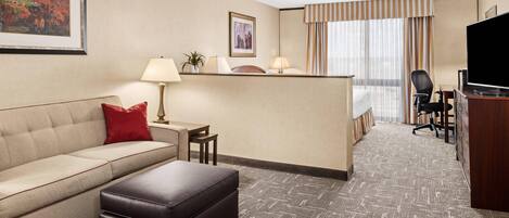Habitación de hotel con sofá, reposapiés, escritorio, lámpara, silla, televisor y cama con cortinas.