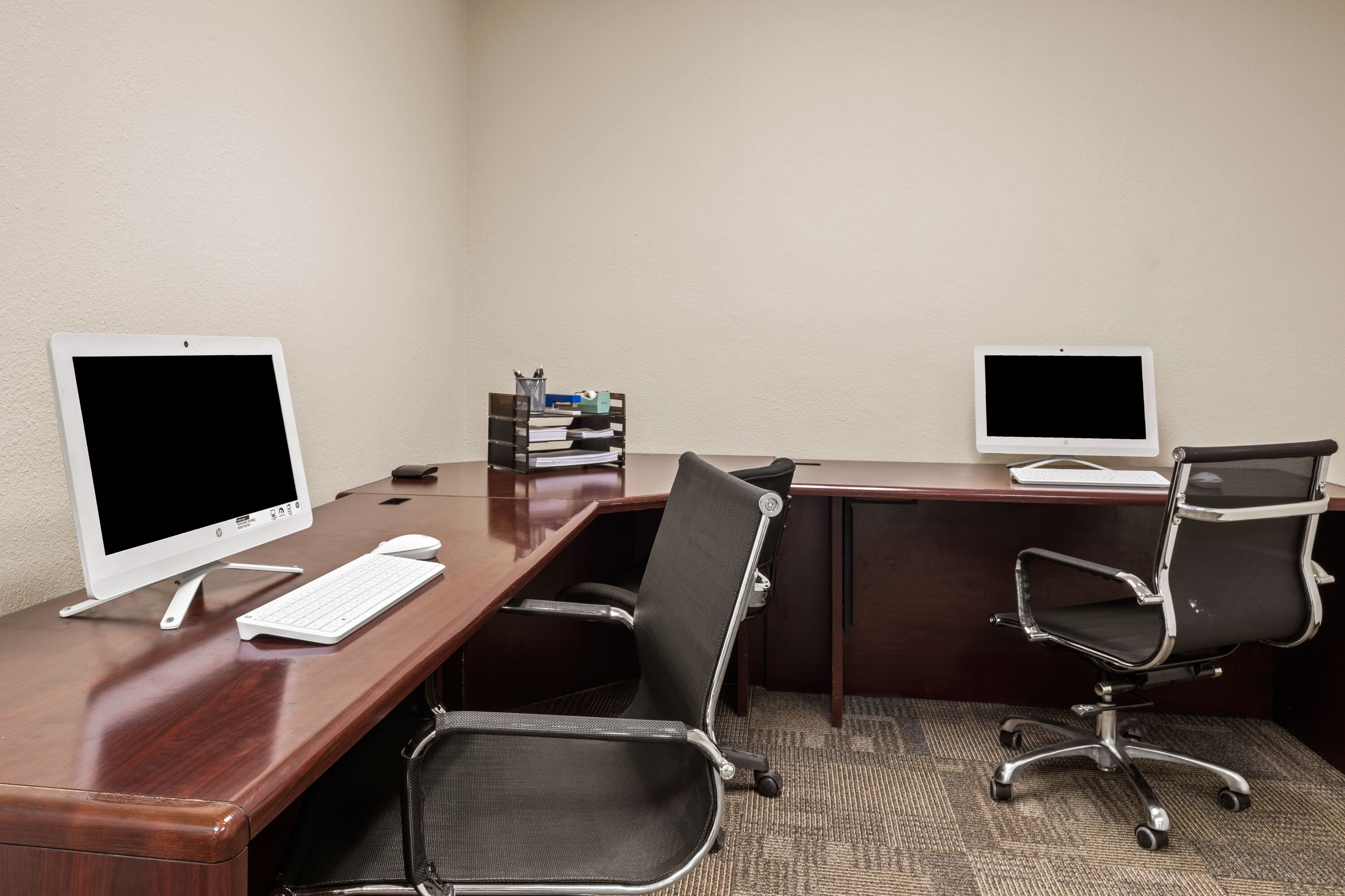 A conference room with two desks, each equipped with a computer monitor, keyboard, and mouse. There are two office chairs, one at each desk. A stack of books and a small container with pens are placed on the central desk.