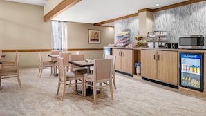 A hotel dining area with a wooden table and chairs, a refrigerator with beverages, a microwave, and a fruit display.