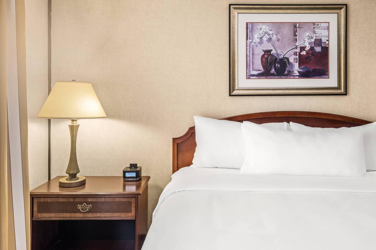 Habitación de hotel con una cama, una mesita de noche con lámpara, un reloj y un cuadro en la pared.