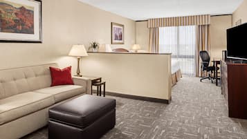 A hotel room with a sofa, ottoman, desk, lamp, television, and a view of the hotel lobby.