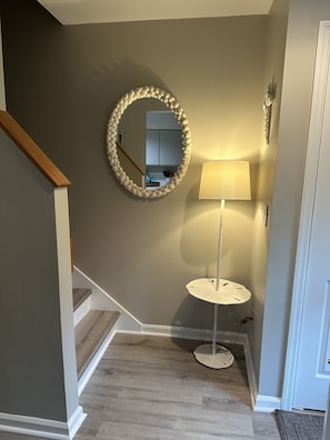 A hallway with a wooden staircase, a round mirror with a textured frame, a floor lamp, and a door. - Hidden Gem on Seabrook Island-visit our fabulous beaches on this magical island! (Seabrook Island)