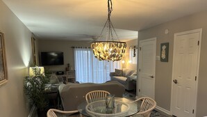 A living room with a glass-top dining table surrounded by wicker chairs, a chandelier, a flat-screen TV, and a sofa.