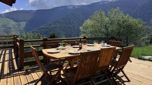 Una terraza de madera con una mesa dispuesta para seis personas, con vistas a un paisaje montañoso.