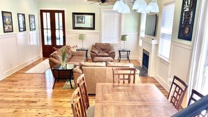 A living room with a brown leather sofa, a glass coffee table, and a wooden dining table with chairs. There are framed pictures on the wall, a fireplace, and a ceiling fan. - Beautiful Cottage on Hilton Head Island, White (Hilton Head Island)