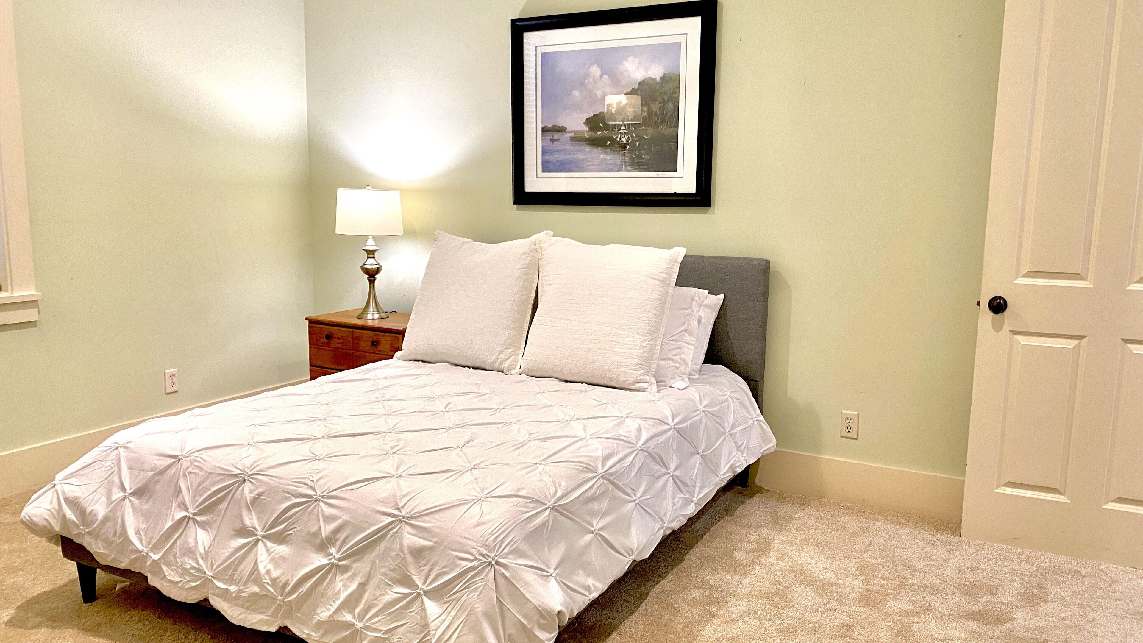 A neatly made bed with a white duvet and pillows, a bedside table with a lamp, and a framed picture on the wall.