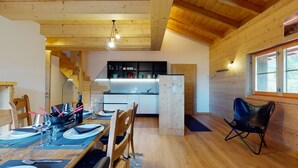 A dining area with a wooden table set for four, a kitchen in the background, and a window with natural light. - Des Alpes (002) - ON THE SLOPES apartment 6 pers (Haute-Nendaz)