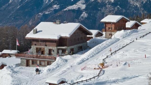 A mountain lodge with a snow-covered roof, wooden exterior, and a ski slope in the foreground.