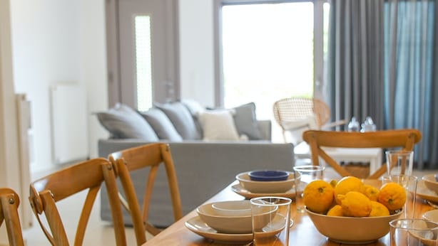 Un comedor con una mesa dispuesta para cuatro, con platos, tazones y vasos. Se observan dos sillas de madera y un sofá al fondo.