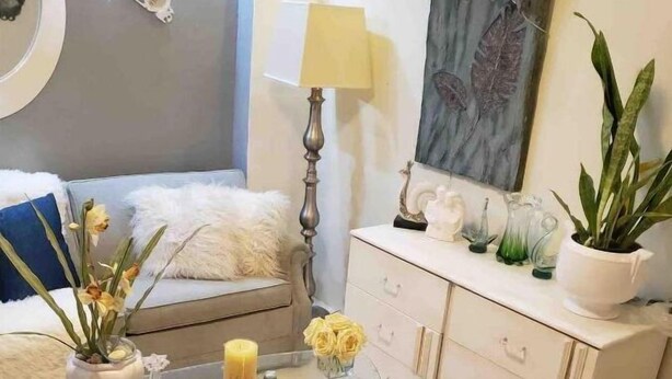 Una sala de estar moderna con una mesa de centro de vidrio, sofá blanco, un taburete blanco mullido y objetos decorativos como flores y una lámpara.