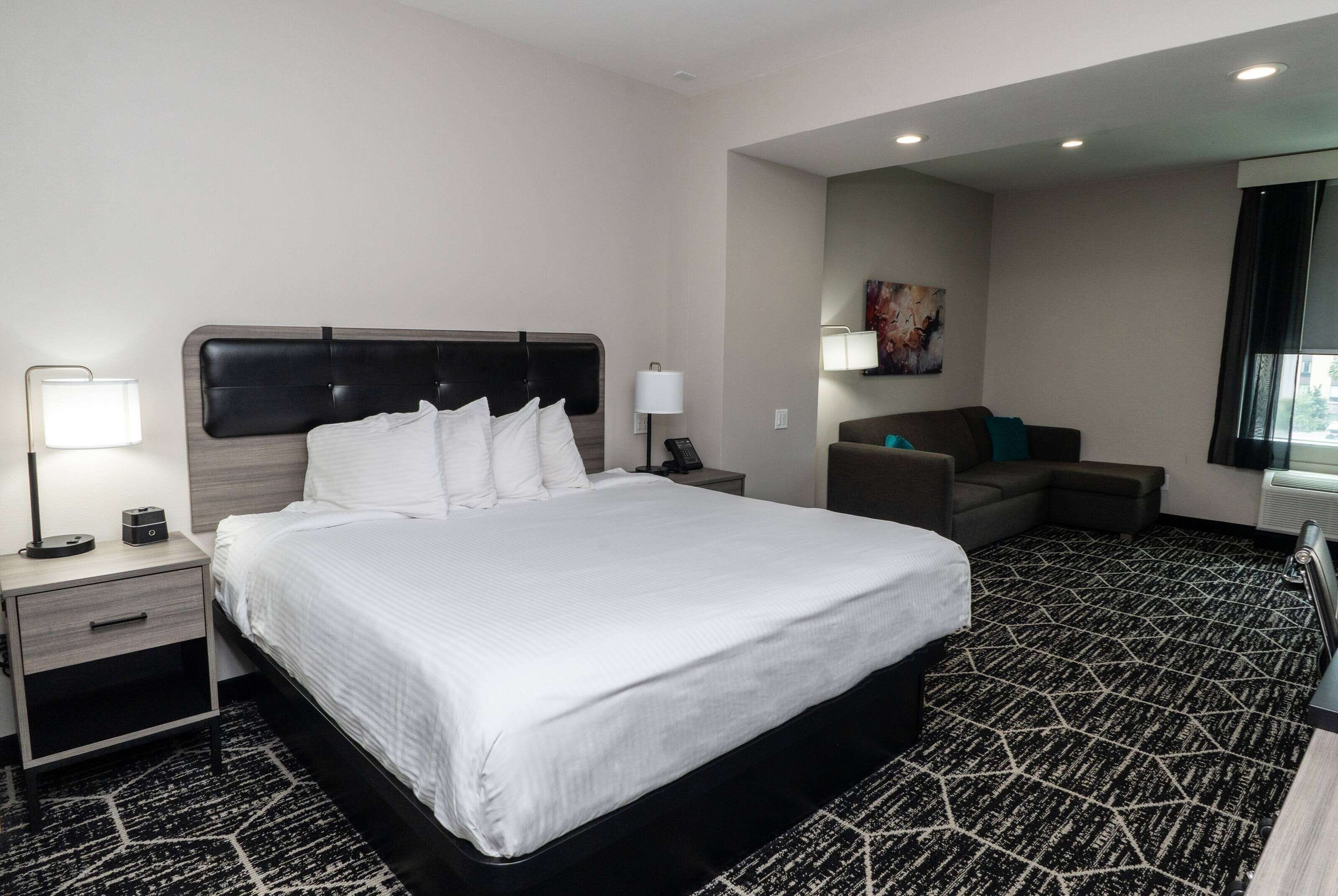 A hotel room with a large bed, a bedside table with a lamp, a small table with a phone, a couch, and a patterned carpet.