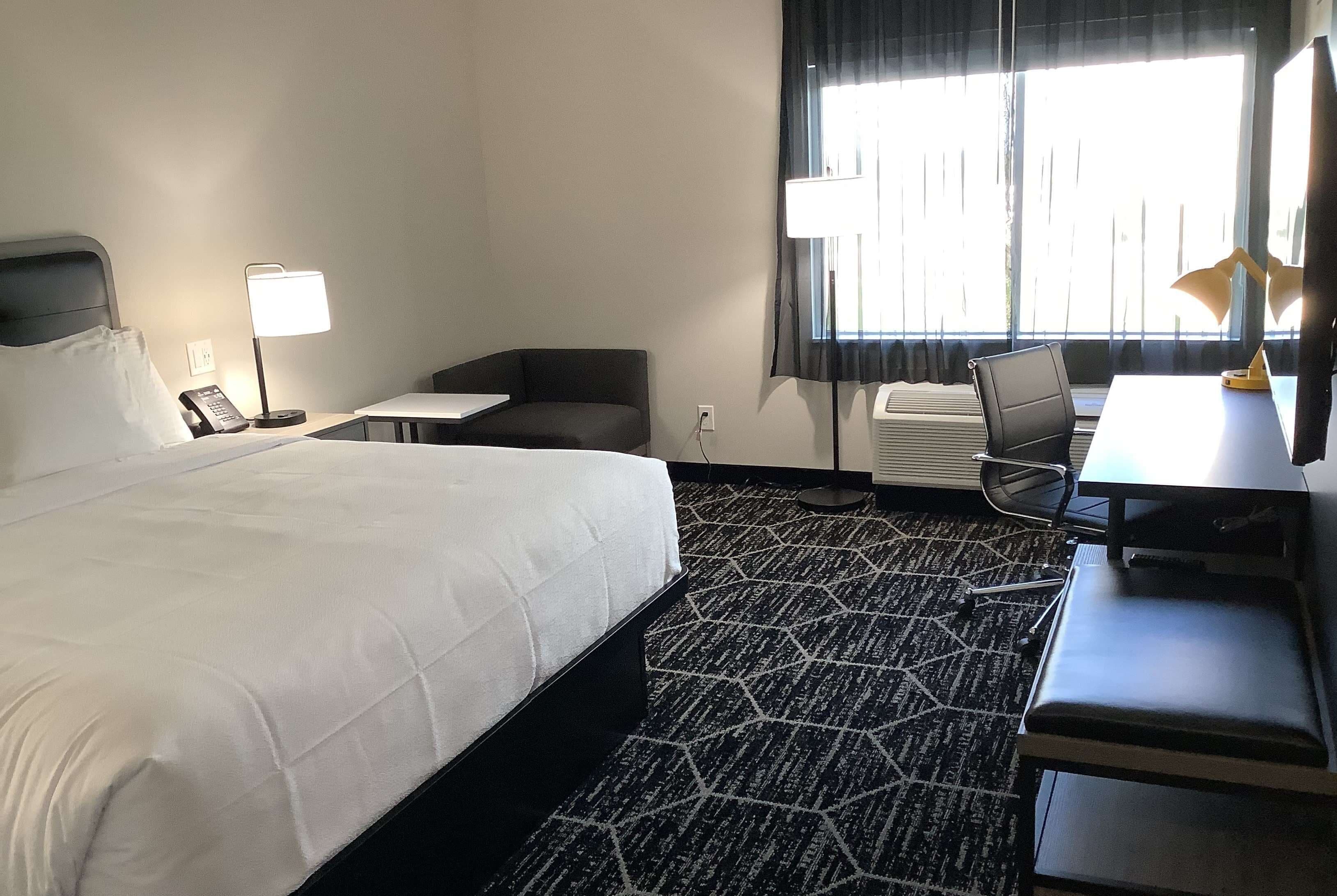 Une chambre d’hôtel avec un grand lit, un bureau avec une chaise, une petite table, un canapé et une fenêtre avec des rideaux.