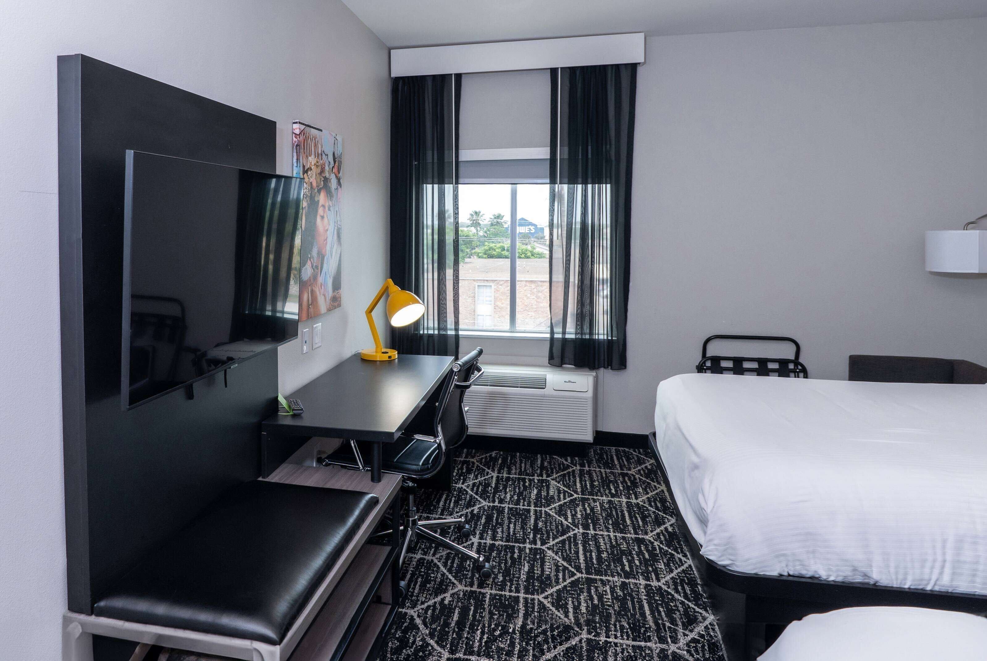 Habitación de hotel con cama, escritorio, silla, televisor y ventana con cortinas.