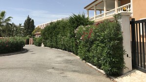 A paved driveway leading to a building with a covered balcony and a black gate. - Location Villa (Fréjus)