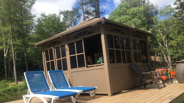A wooden deck with two blue lounge chairs and a small wooden structure with windows. - Tiny House on the River with Wifi, Sauna and Hot Tub. (Burks Falls)