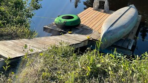 A wooden dock with a green tire and a boat, surrounded by lush greenery and a calm river. - Tiny House on the River with Wifi, Sauna and Hot Tub. (Burks Falls)