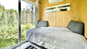 A bedroom with a large window, wooden walls, a bed with grey bedding, and an air conditioning unit. - Tiny House on the River with Wifi, Sauna and Hot Tub. (Burks Falls)
