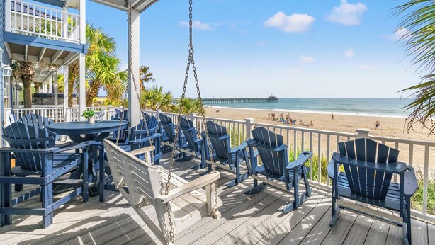 A wooden deck with blue chairs and a swing, overlooking a beach with a pier in the distance.