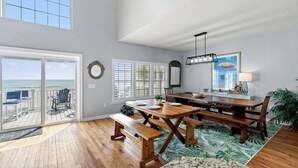 A spacious dining area with a large wooden table and benches, a view of the ocean through sliding glass doors, and a ceiling light fixture.