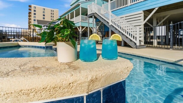 A pool area with a blue building in the background, a potted plant, and two drinks with lemon slices.