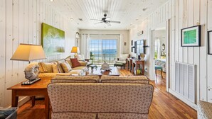 A spacious living room with a large window, a ceiling fan, and a view of the ocean. - Casablanca - Luxury Oceanfront Home in Cherry Grove with Game Room & Pool (North Myrtle Beach)