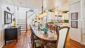 A spacious dining area with a wooden table and chairs, a staircase leading to an upper level, and a kitchen with white cabinets and green appliances. - Casablanca - Luxury Oceanfront Home in Cherry Grove with Game Room & Pool (North Myrtle Beach)