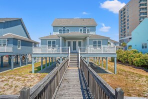 A beachfront property with a wooden boardwalk leading to a two-story house with a balcony. - Cassie - Pet Friendly, Oceanfront home in Cherry Grove (North Myrtle Beach)