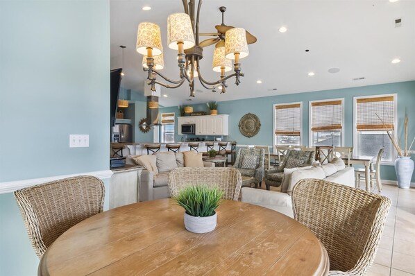 A dining area with a round wooden table, wicker chairs, and a chandelier. - Tropical Pleasure, Luxury Oceanfront 6 Bedroom Home in Cherry Grove - 3 Large Balconies, Game Room, Pool & Hot Tub (North Myrtle Beach)