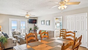 A dining area with a wooden table, woven placemats, and wooden chairs. There are ceiling fans, a television, and a view of the outdoors through a door. - Chase - Pet Friendly Oceanfront House in Cherry Grove (North Myrtle Beach)