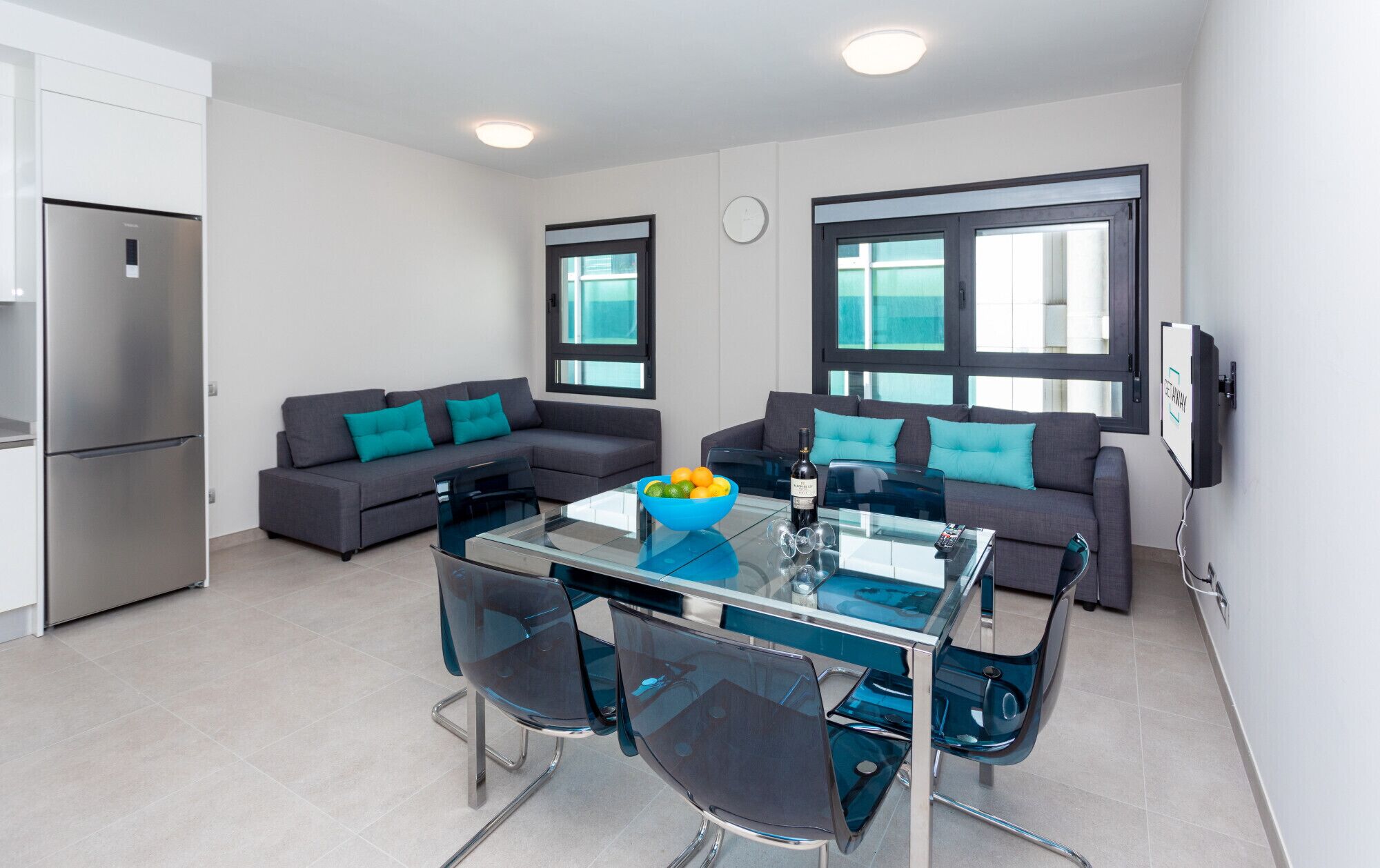 A modern kitchen with a built-in refrigerator, a glass dining table with chairs, and two grey sofas with teal cushions.