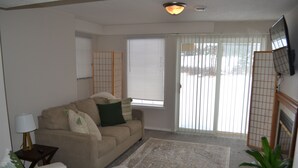 Una sala de estar con un sofá beige, un sillón blanco, una mesita con una lámpara y un ventanal con persianas.