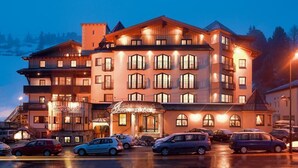 A multi-story hotel with a traditional architectural style, featuring a central entrance with a sign, multiple balconies, and a parking area with several cars. - Tauernkoenig (Tweng)