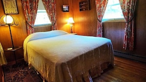 A neatly made bed in a room with wooden walls, a small bedside table with a lamp, and floral curtains. - Eagle Landing, Views, WI-FI, Private, Spacious! (Boone)