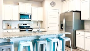 Una cocina con armarios blancos, un refrigerador de acero inoxidable, un microondas y un lavamanos. Hay tres taburetes azules frente a la encimera.