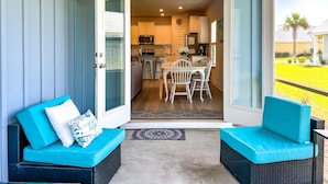 Un espacio al aire libre techado con una mesa y sillas, una cocina al fondo y dos zonas de estar con cojines azules.