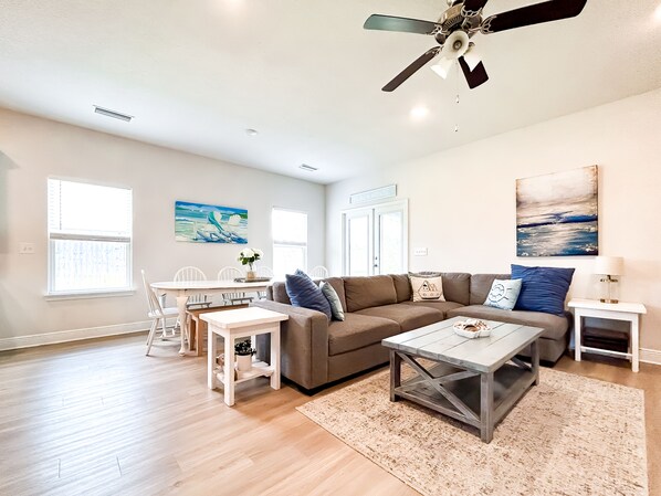 A spacious living room with a ceiling fan, a dining area with a table and chairs, and a large window.