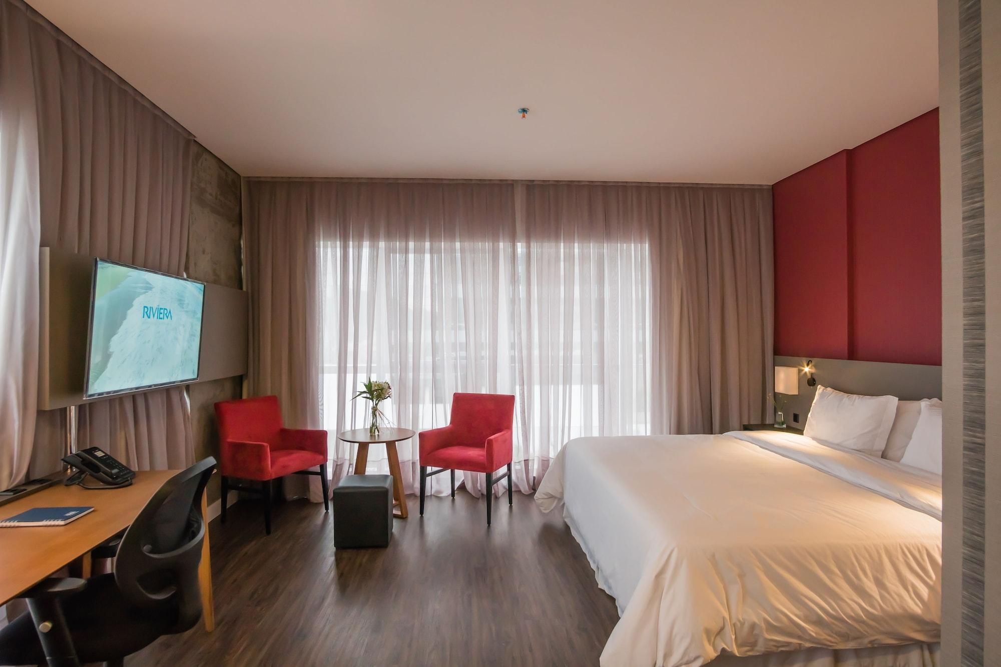 Una habitación de hotel con cama, escritorio con teléfono, dos sillas rojas, una mesita con un florero y un televisor en la pared.