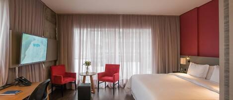 Una habitación de hotel con cama, escritorio con teléfono, dos sillas rojas, una mesita con un florero y un televisor en la pared.
