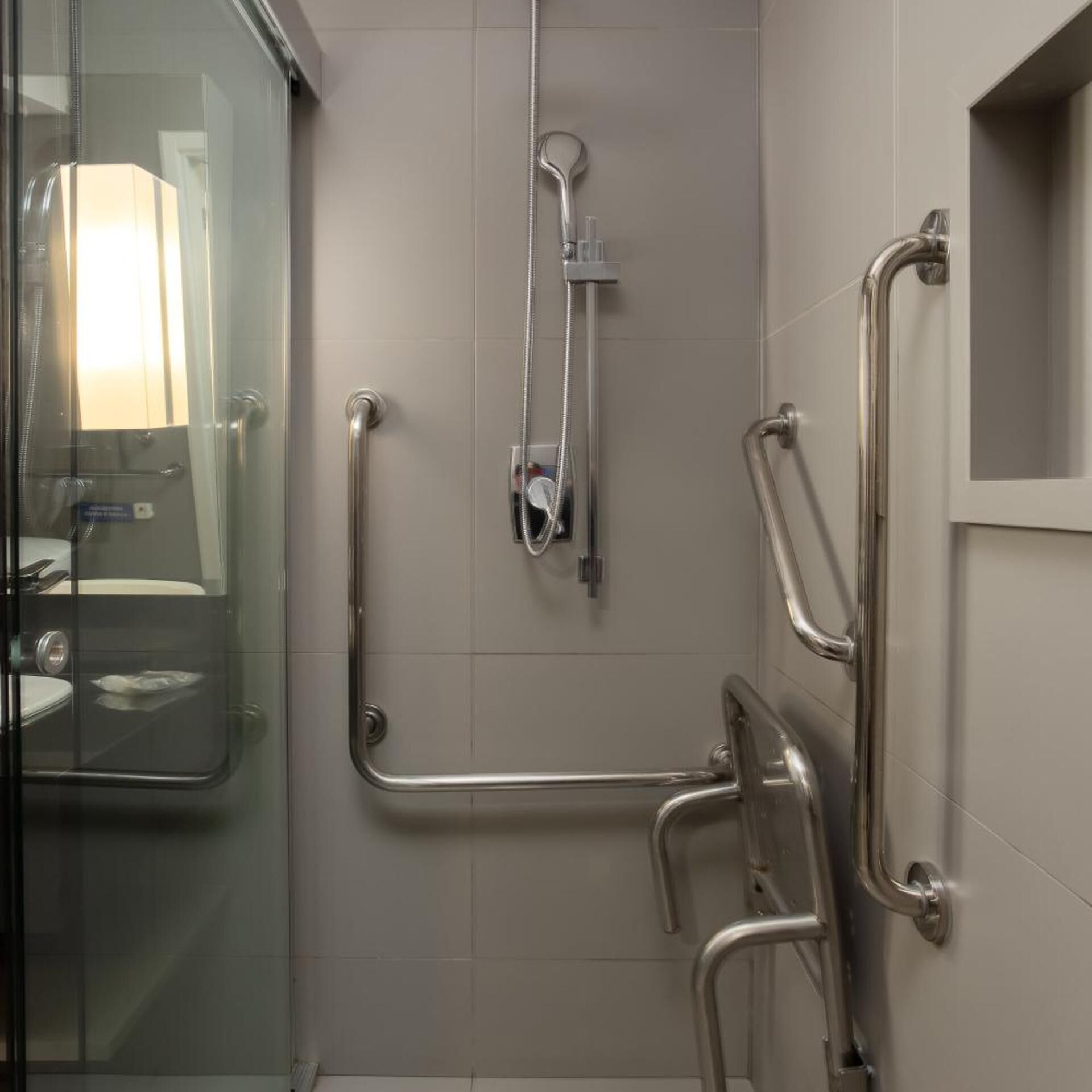 A bathroom with a glass partition, a shower with a handheld showerhead, and a handrail.