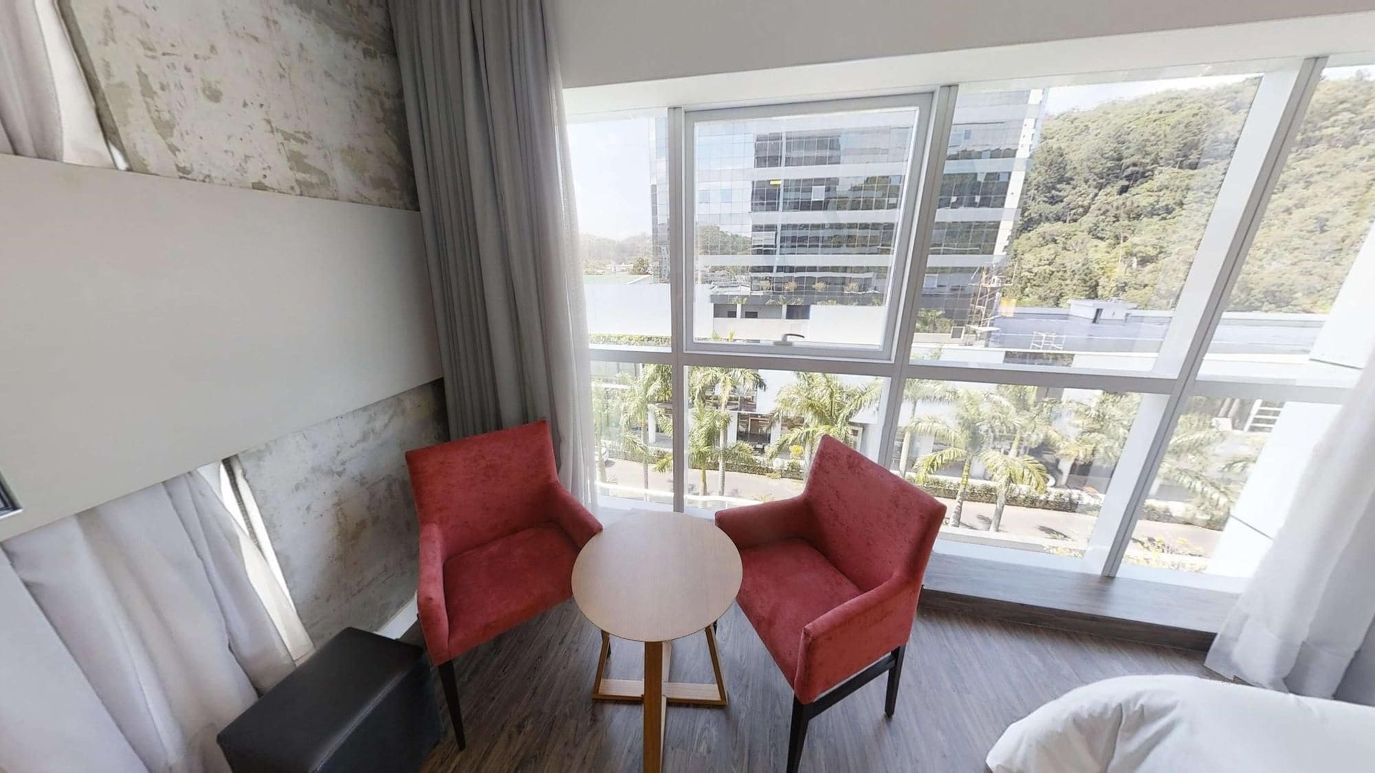 A hotel room with a large window, two red chairs, a small round table, and a white bed.