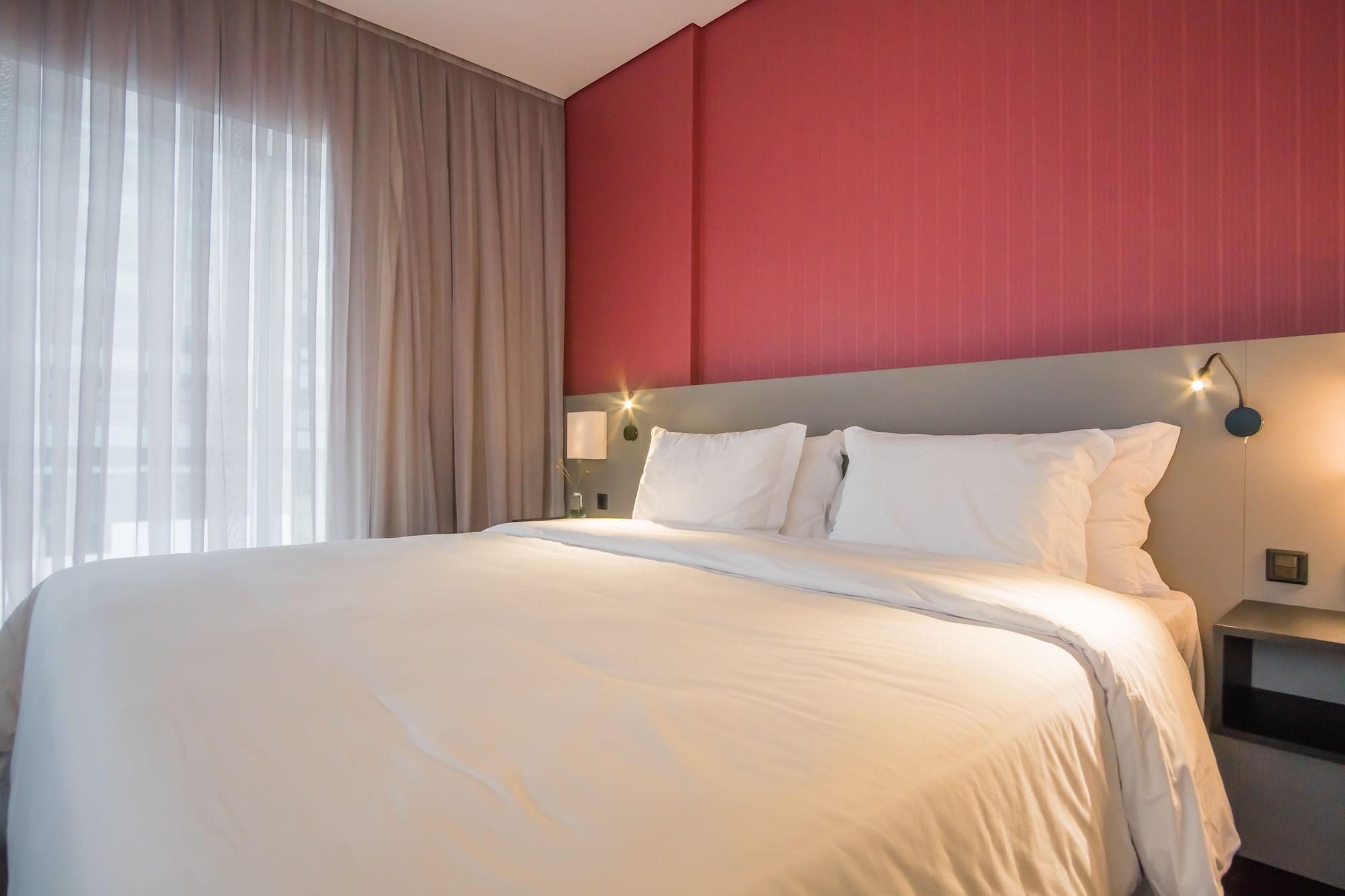 Una habitación de hotel con una cama grande, una pared de acento roja y una ventana con cortinas traslúcidas.