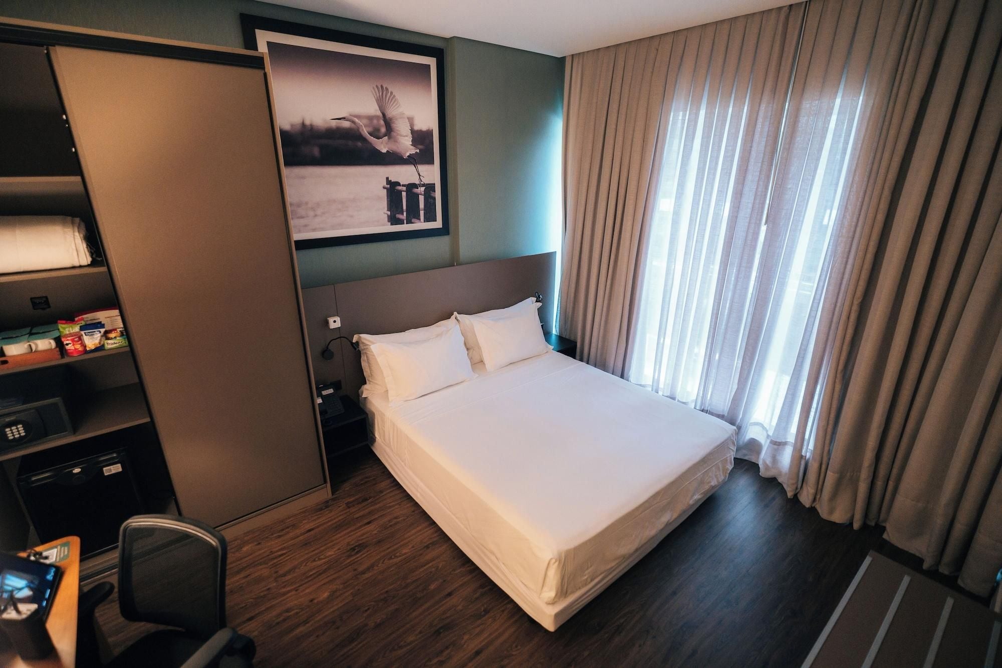 A hotel room with a bed, a desk with a chair, a wardrobe, and a window with curtains.