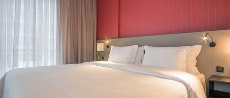 Una habitación de hotel con una cama grande, una pared de acento roja y una ventana con cortinas traslúcidas.