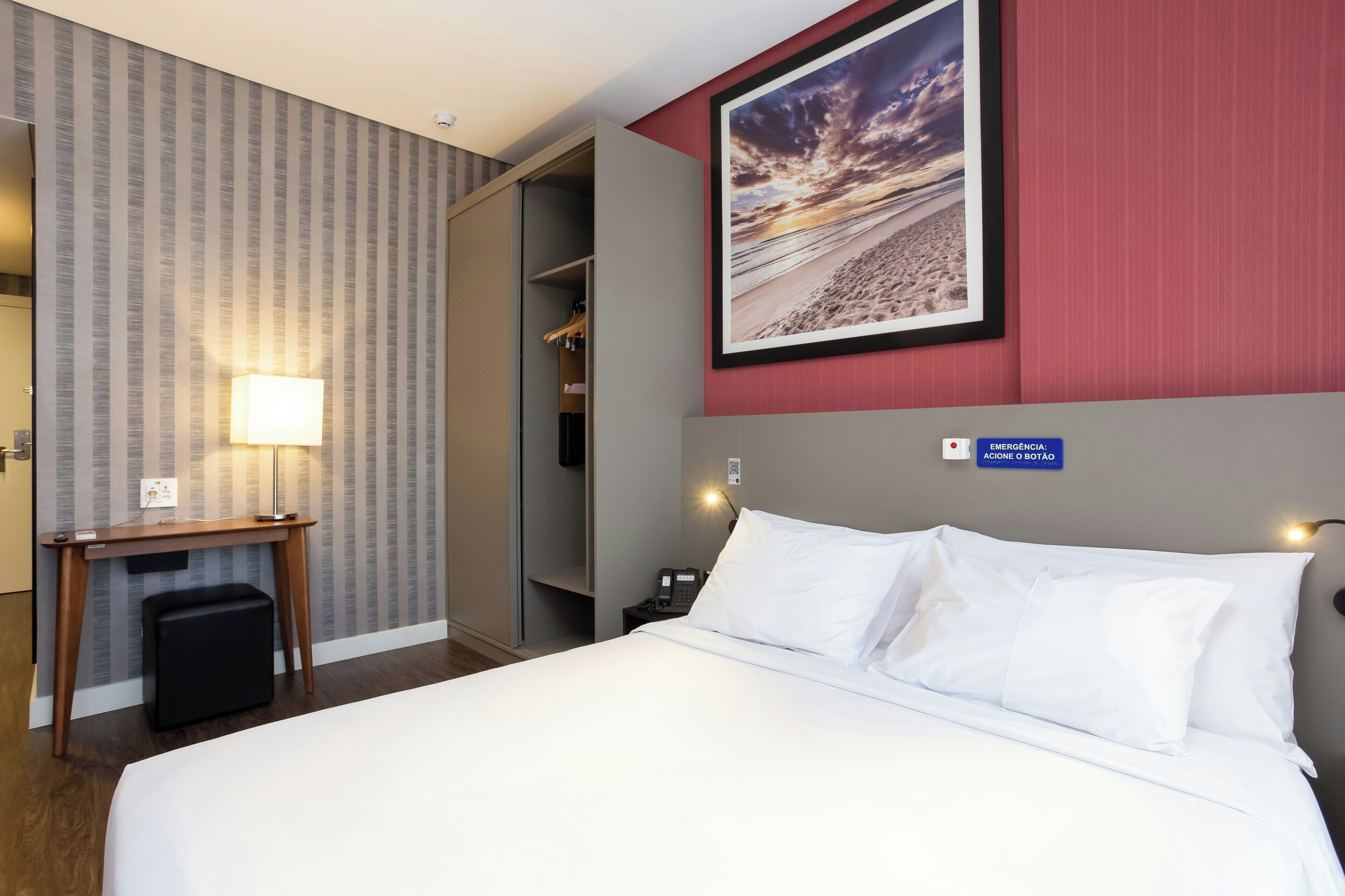 A hotel room with a bed, a desk, a lamp, a wardrobe, and a framed picture on the wall.